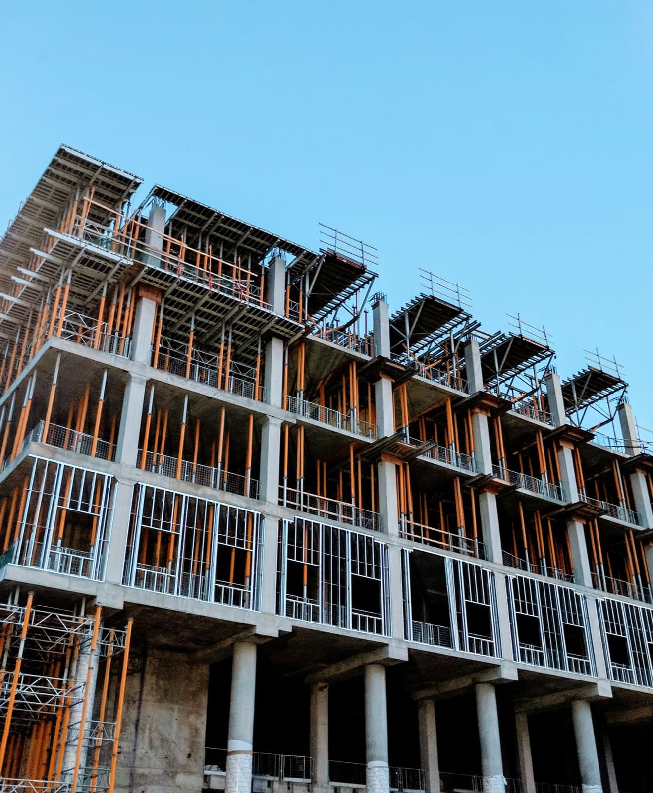 The concrete frame of a multi-storey building under construction, clad in scaffolding against a clear sky.