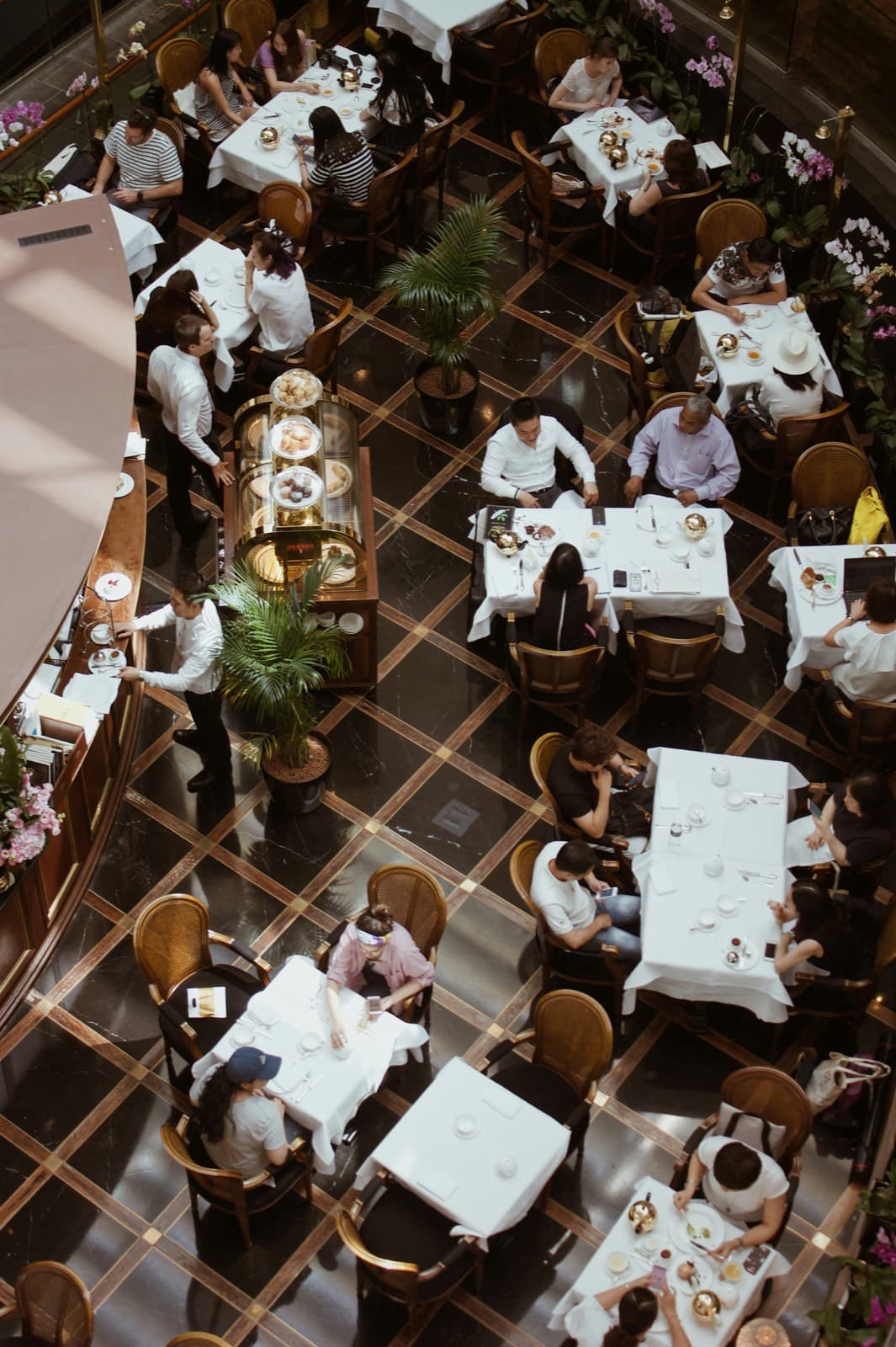 A busy restaurant floor seen from above, with white-linen tables and waiting staff between them.