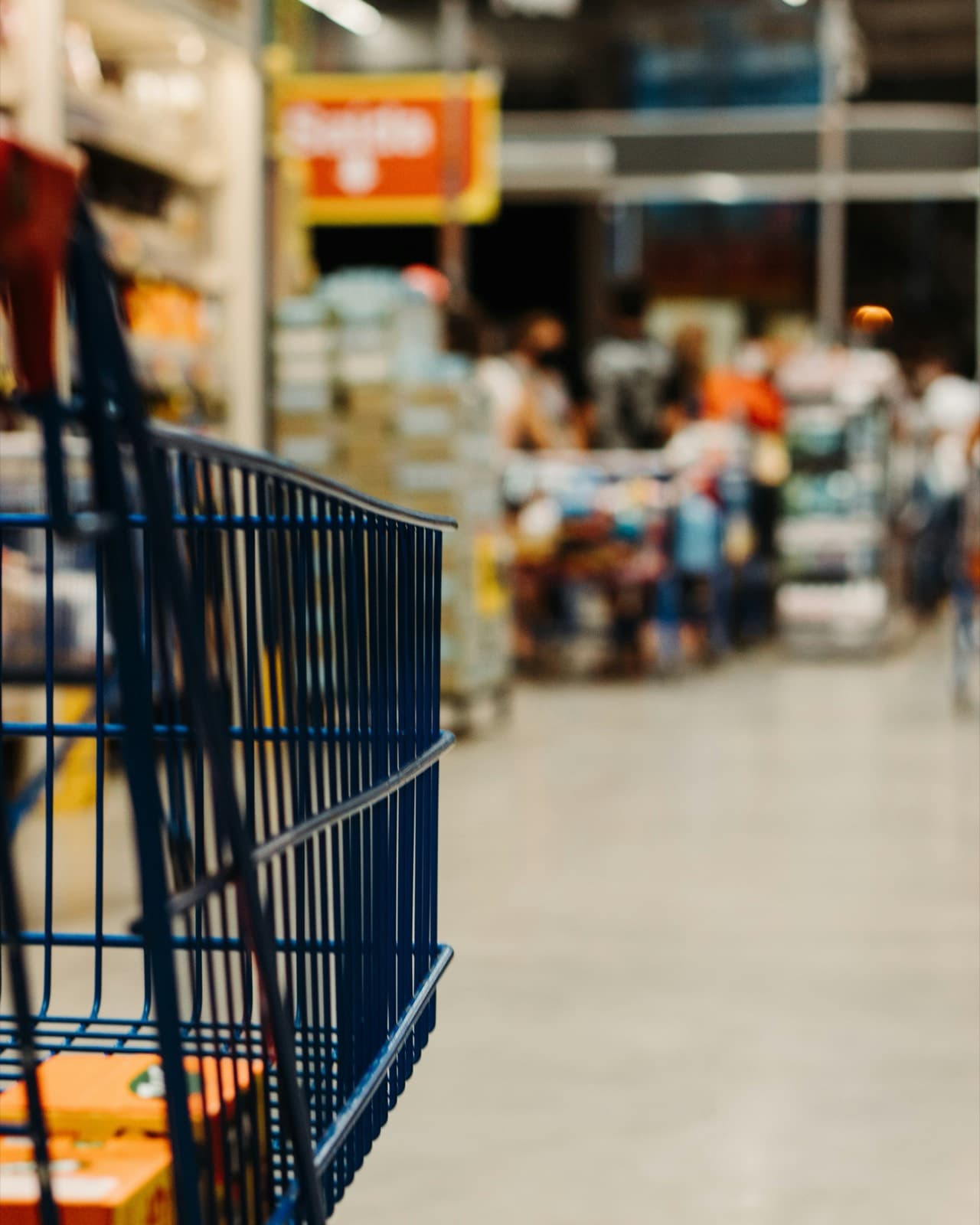 A trolley in the foreground of a warehouse store aisle, shoppers blurred beyond it.